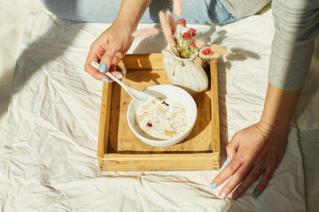 Woman in jeans sitting on the bed, and eating healthy granola bowl during the morning sunlight, breakfast in the bed.