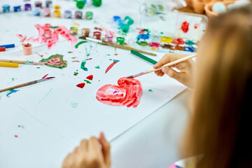 Cute child sitting at the table and drawing red heart on white paper