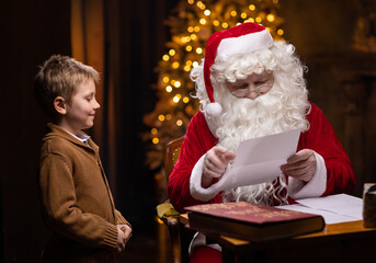 Santa Claus and little boy. Cheerful Santa is working while sitting at the table. Fireplace and Christmas Tree in the background. Christmas concept.