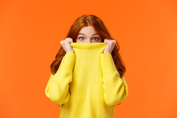 Surprised and astonished cute curious redhead female pulling sweater collar on face and popping...