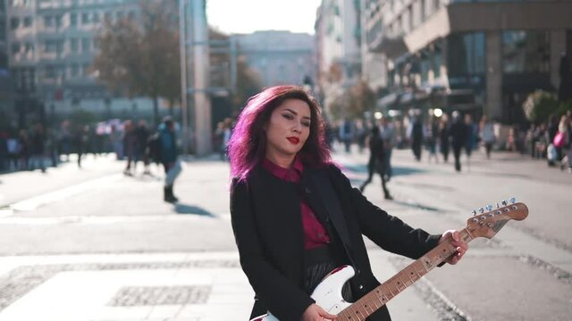 Music lifestyle. Daring rebel woman with purple hair playing electric guitar at city center during bright sunset. People walking in background. Asian girl like rock star performing show at the street.