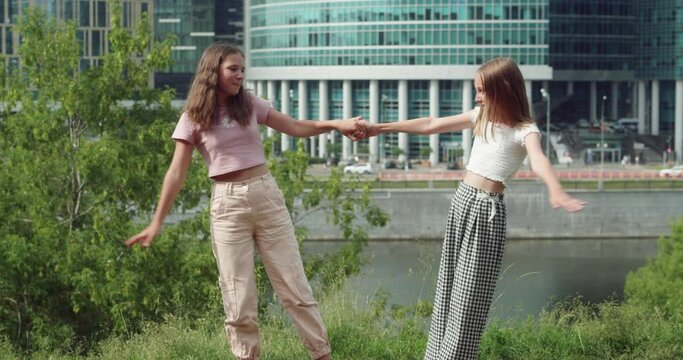 Happy young girls holding hands on embankment in city. Cute smiling teenage girls having fun together with modern high rise buildings on background
