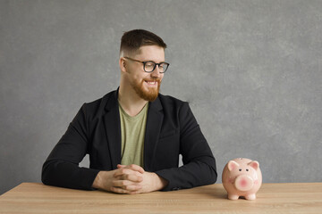 Cheerful handsome caucasian man sitting at the table and looking at the piggy bank with a smile on...