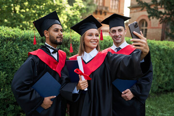 Portrait of three young international graduate student friends making selfie in graduation robes in campus