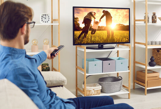Man watching movie on television at home. Relaxed guy sitting on couch in living room in front of big TV screen, watching film, series, serial or collection of photo memories of family summer holiday