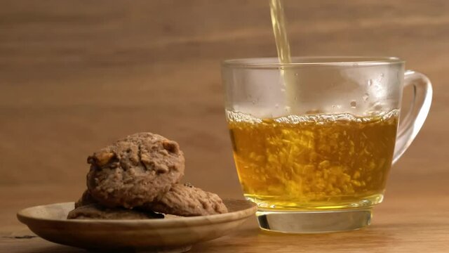 Pouring hot tea into transparent glass with pile of delicious homemade chocolate chip butter cookies in wooden plate on wooden table.