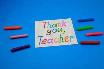 Greeting handwritten card with words of gratitude to the teacher, dedicated to the teacher's day, colored crayons lie on a blue background