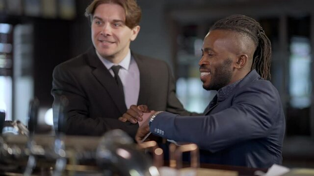 Handsome positive African American man in suit texting on smartphone greeting Caucasian colleague entering bar. Portrait of two positive coworkers resting after work indoors. Business friendship