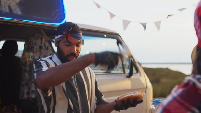 Young positive middle eastern seller in disposable gloves cooking hot dog and chatting with people while working at food truck on summer festival