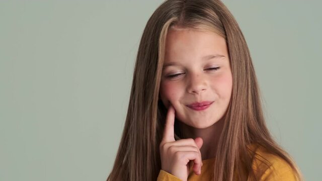 A close-up view of a thinking small girl standing isolated over gray wall in the studio