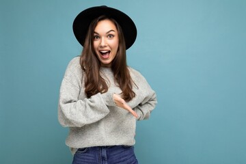 Photo of beautiful brunette happy joyful young woman directing finger to the side with copy space, demonstrating and presenting low prices poster, wearing gray sweater and black hat, isolated on blue