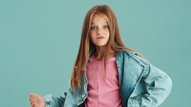 A strict small girl wearing jeans jacket shaking her finger while standing isolated over a blue wall in the studio