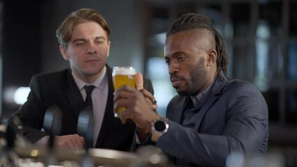 African American beer connoisseur talking with Caucasian friend explaining pale lager features pointing at glass. Portrait of concentrated men in formal suits resting in bar after work indoors
