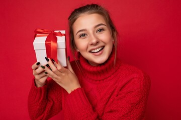 Naklejka premium Shot of attractive positive smiling young brunette woman isolated over colourful background wall wearing everyday trendy outfit holding gift box and looking at camera