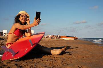 Portrait of beautiful female surfer with her surfboard. Young woman having video call while relaxing at the beach...