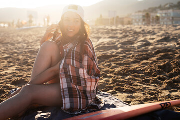 Fototapeta premium Portrait of beautiful female surfer with her surfboard. Young woman talking to the phone while relaxing at the beach...