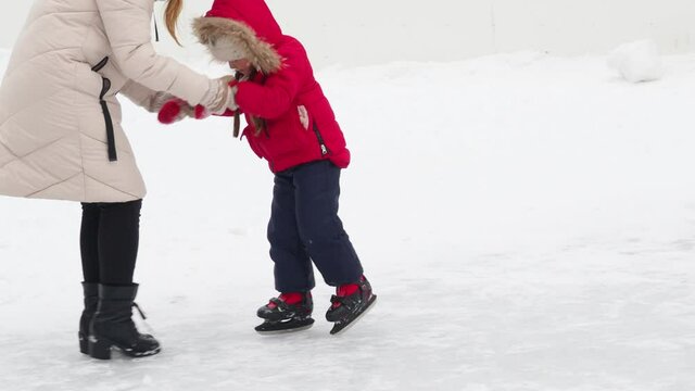 family in winter skating on an open skating rink on ice preschooler boy and girl learn to skate on ice rink. Kid wearing winter jacket, hood hat and neckwarmer standing on one knee mom teaches to ridу