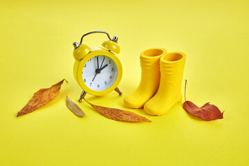 Fall Back Daylight Saving Time concept with yellow clock, boots and autumn leaves on yellow background