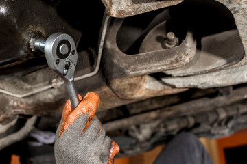 A mechanic unscrews an oil drain plug underneath the chassis of a SUV with a ratchet wrench. First...
