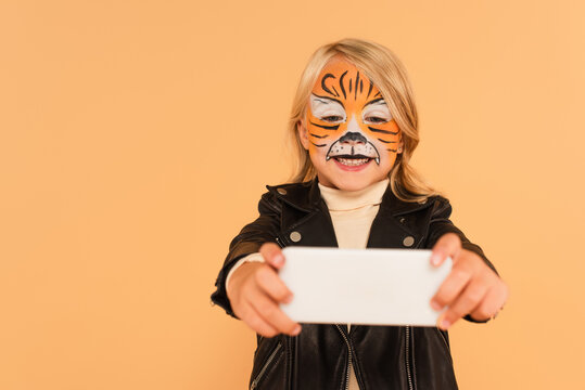 smiling girl in tiger makeup taking selfie on blurred smartphone isolated on beige