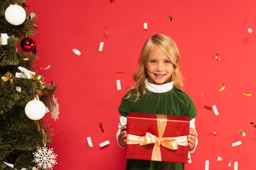 happy kid with gift box smiling at camera near christmas tree under falling confetti isolated on red