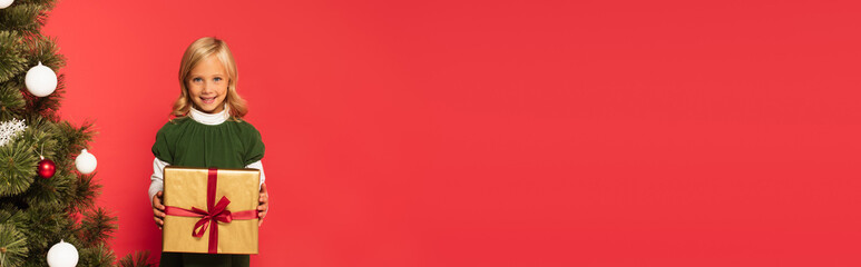 happy girl holding present near decorated christmas tree isolated on red, banner