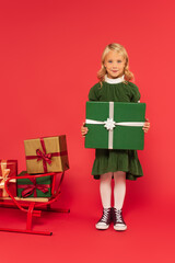 positive girl holding green gift box near sled with presents on red