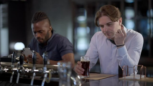 Portrait of drunken Caucasian man sitting in bar with dark lager beer and car keys as blurred African American friend ordering taxi online in smartphone app. Responsible driving and booze