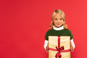 smiling child holding present in golden wrapping paper while looking at camera isolated on red