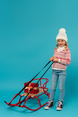 Fototapeta na wymiar full length view of kid in warm sweater and jeans smiling near sled with gift box on blue
