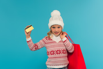 kid in knitted sweater and hat standing with credit card and shopping bag isolated on blue