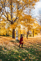 Obraz premium Rear view of elegant young woman in a burgundy casual coat in autumn. Cute model walks in the park in golden autumn through colorful trees and fallen leaves. Autumn walk, colorful nature.