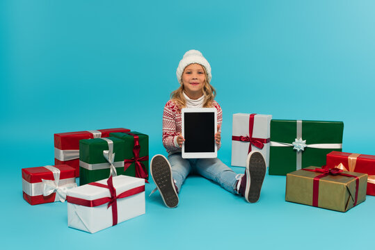 happy kid in jumper and hat sitting with digital tablet near multicolored gift boxes on blue
