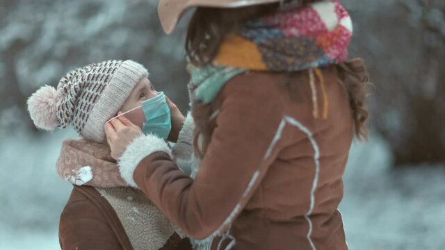 stylish mother and daughter in a hats and sheepskin coats with medical mask wearing medical mask outdoors in the city park in winter.