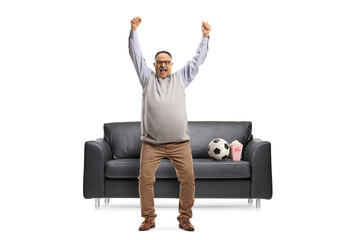 Full length portrait of a happy mature football fan cheering in front of a sofa
