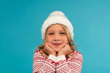 happy child in knitted hat and warm sweater with ornament looking at camera isolated on blue