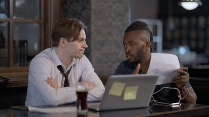 Focused Caucasian and African American men brainstorming sitting in restaurant indoors. Portrait of busy concentrated managers talking discussing strategy idea. Teamwork and lifestyle