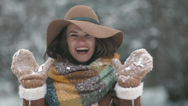 happy stylish middle aged woman in brown hat and scarf with snowy mittens in sheepskin coat outdoors in the city park in winter.