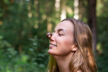 A snail sits on the nose of a Caucasian woman. The girl laughs and looks at the clam. Close-up portrait against the background of a green forest