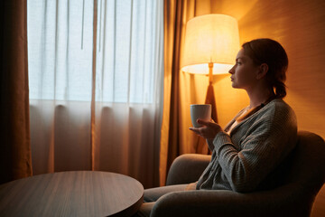 A young woman is sitting in a room near the window. She has a cup in her hands. Evening, the room is flooded with soft light from a floor lamp. Home comfort, rest. Hotel. Quarantine, self-isolation.