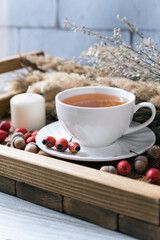 White cup of tea with saucer, rosehip berries, acorns, candles and dried plants on a small wooden tray. Vertical photo