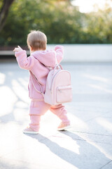 Funny baby girl 1 year old wear stylish pink sport suit and backpack walk on street over urban background. Cute little child with trendy clothes. Childhood. Back view.