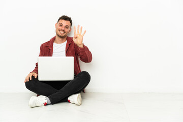 Young handsome caucasian man sit-in on the floor with laptop happy and counting four with fingers