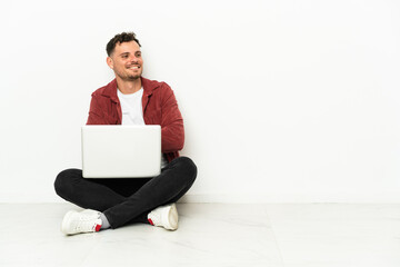 Young handsome caucasian man sit-in on the floor with laptop with arms crossed and happy