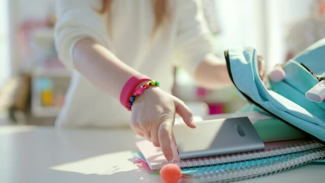 Closeup on school girl with tablet PC, workbooks and blue backpack packing for school at home in sunny day.