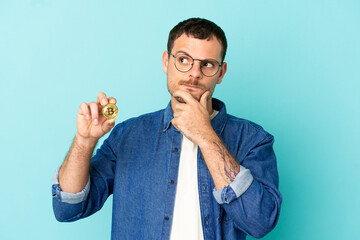 Brazilian man holding a Bitcoin over isolated blue background having doubts and thinking