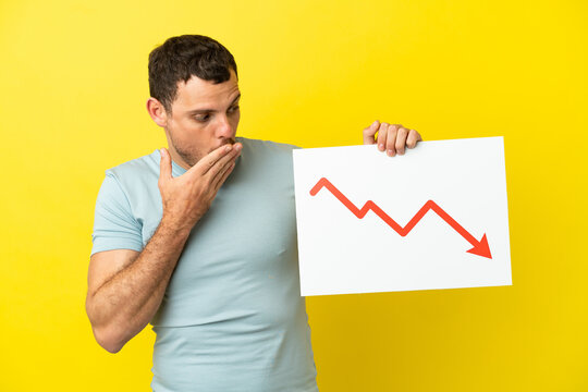 Brazilian man over isolated purple background holding a sign with a decreasing statistics arrow symbol with surprised expression