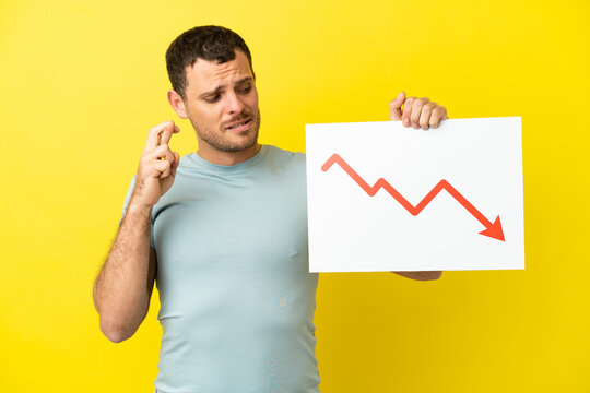 Brazilian man over isolated purple background holding a sign with a decreasing statistics arrow symbol with fingers crossing