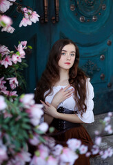 Obraz na płótnie Canvas Beautiful long haired girl in medieval dress sitting on steps in front of blue door surrounded by magnolia flowers