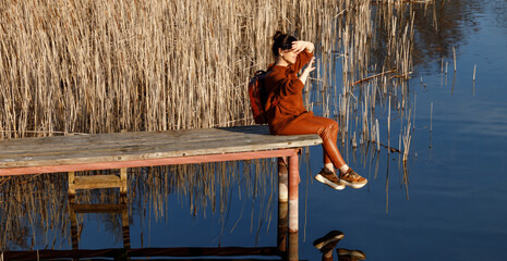 Portrait of a woman in autumn cozy orange clothes with backpack sitting on a wooden bridge near the lake with blue water and looking agains the sun. People, travel, nature concept.